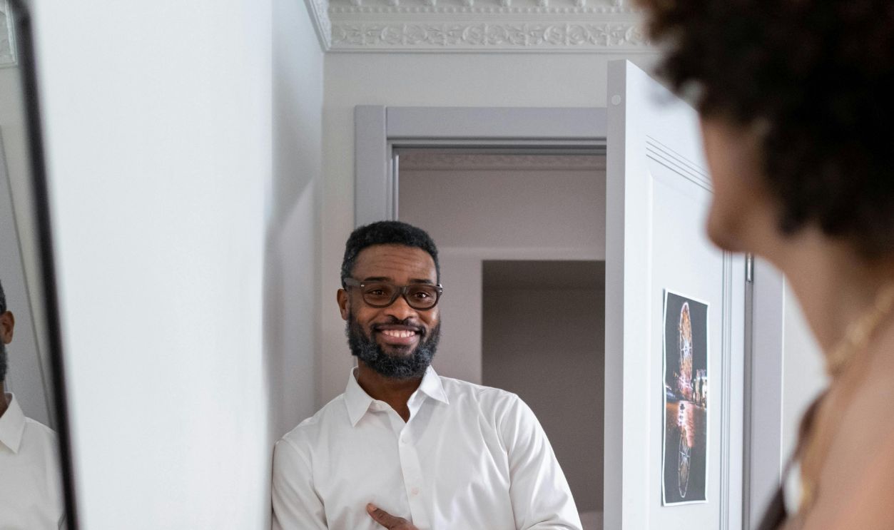 A smiling man in smart casual attire stands indoors by an open door, sharing a joyful moment.