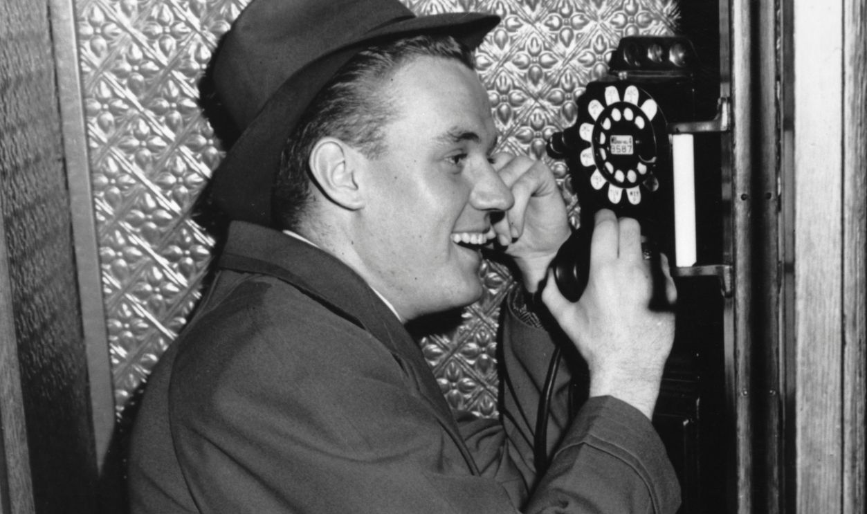 Hanging On The Telephone A smiling man in a fedora and an overcoat holding the receiver as he makes a telephone call from a payphone, United States, circa 1955.