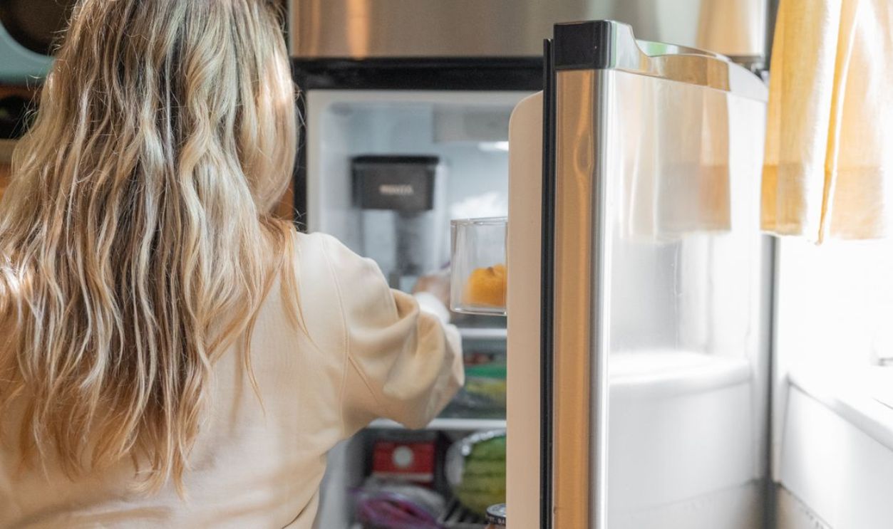 Back view of a blonde woman retrieving food from a refrigerator in a bright kitchen