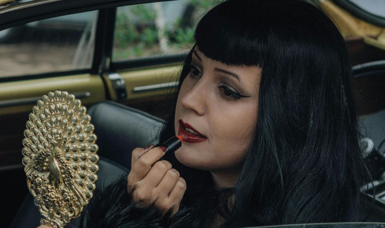 A woman in a vintage car applies red lipstick using an ornate handheld mirror