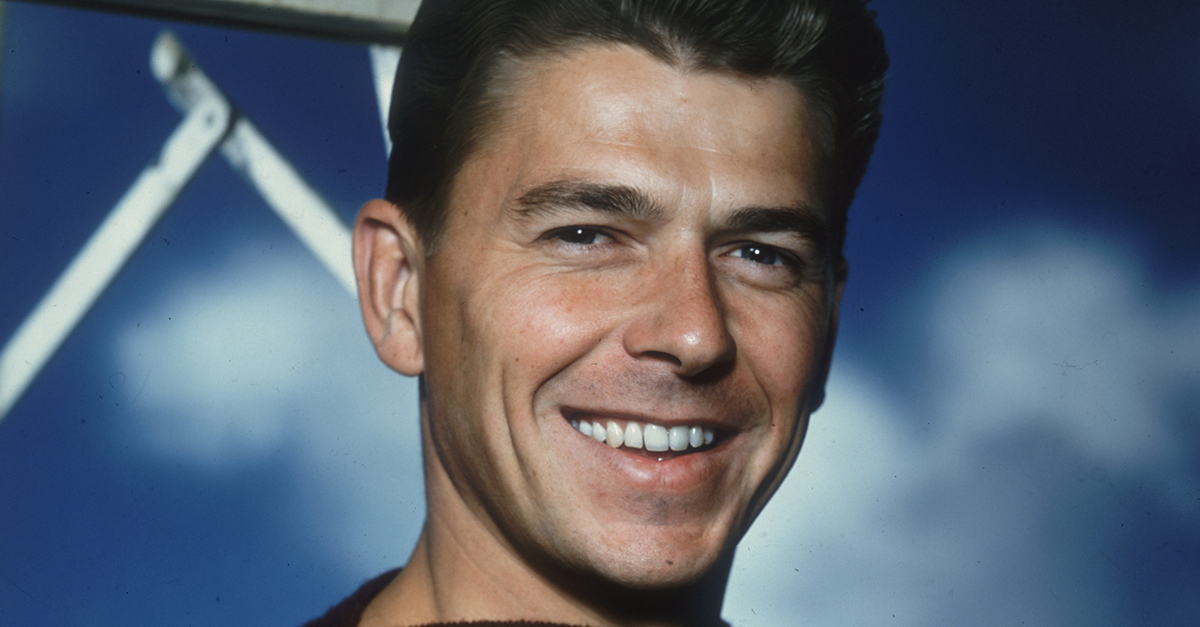 Studio portrait of American actor Ronald Reagan, wearing a short sleeve boat-neck sweater, standing near a ladder against a blue sky backdrop.