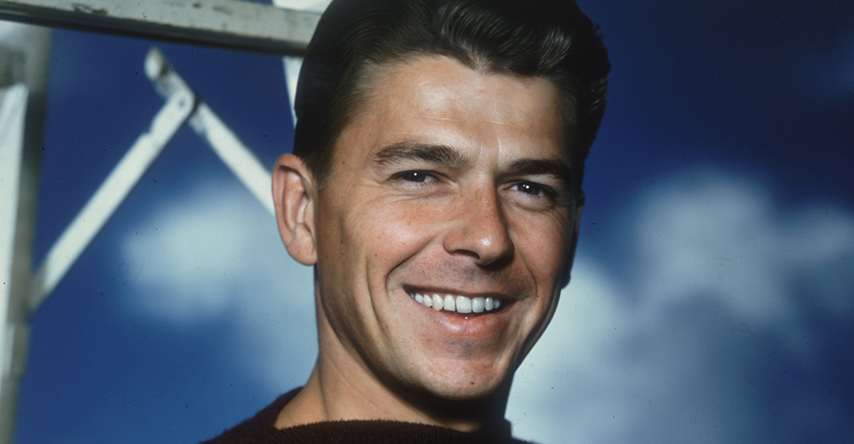 Studio portrait of American actor Ronald Reagan, wearing a short sleeve boat-neck sweater, standing near a ladder against a blue sky backdrop.