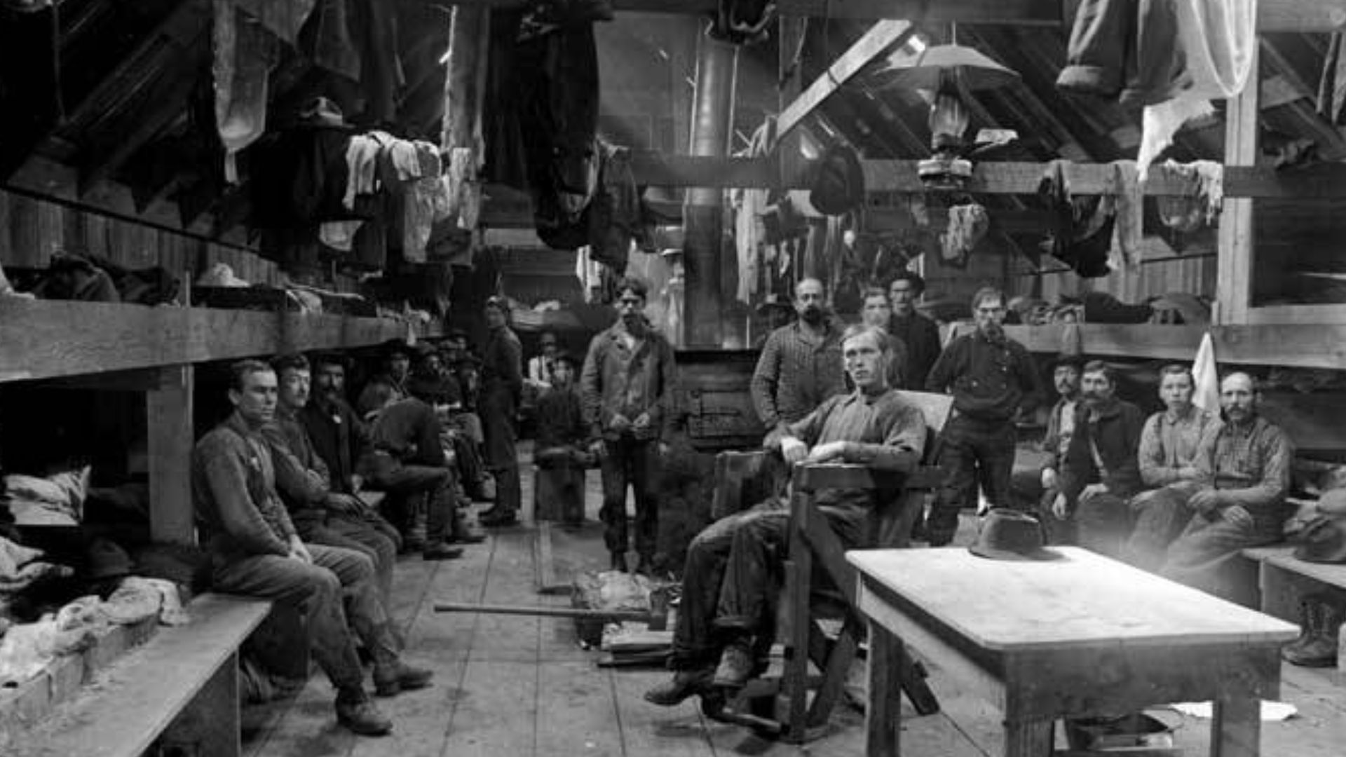 The men who worked in logging camps lived together in crude bunkhouses. Conditions in the camps were harsh, uncomfortable, and dirty.
This photo was taken at a logging camp somewhere in Oregon or Washington. Many of the men are seated at the edge of their