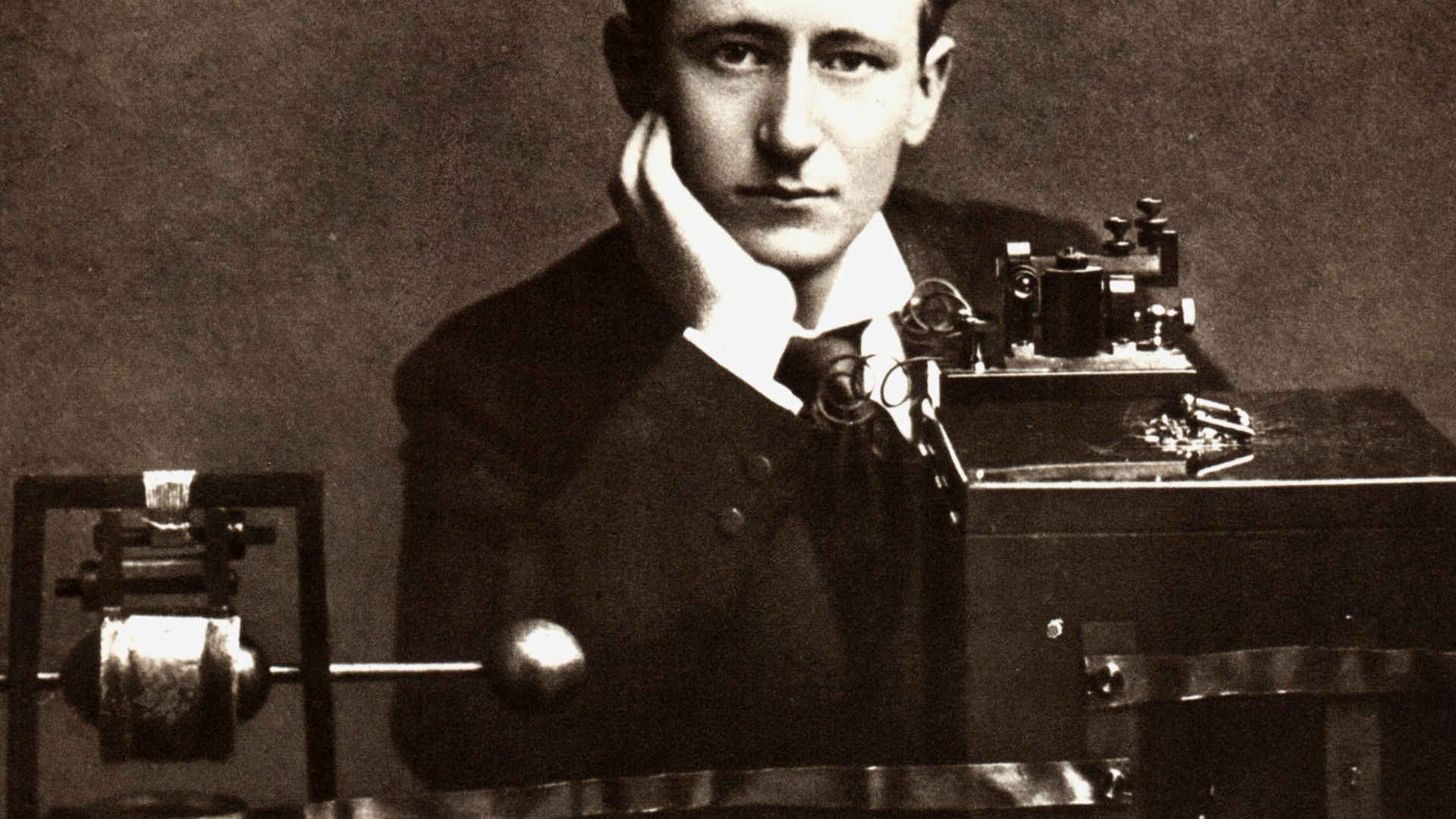 A publicity photo of Italian radio pioneer Guglielmo Marconi posing in front of his early radio apparatus.  The device at left is the transmitter, consisting of a Righi spark gap dipole antenna (balls) which is powered by high voltage from an induction co