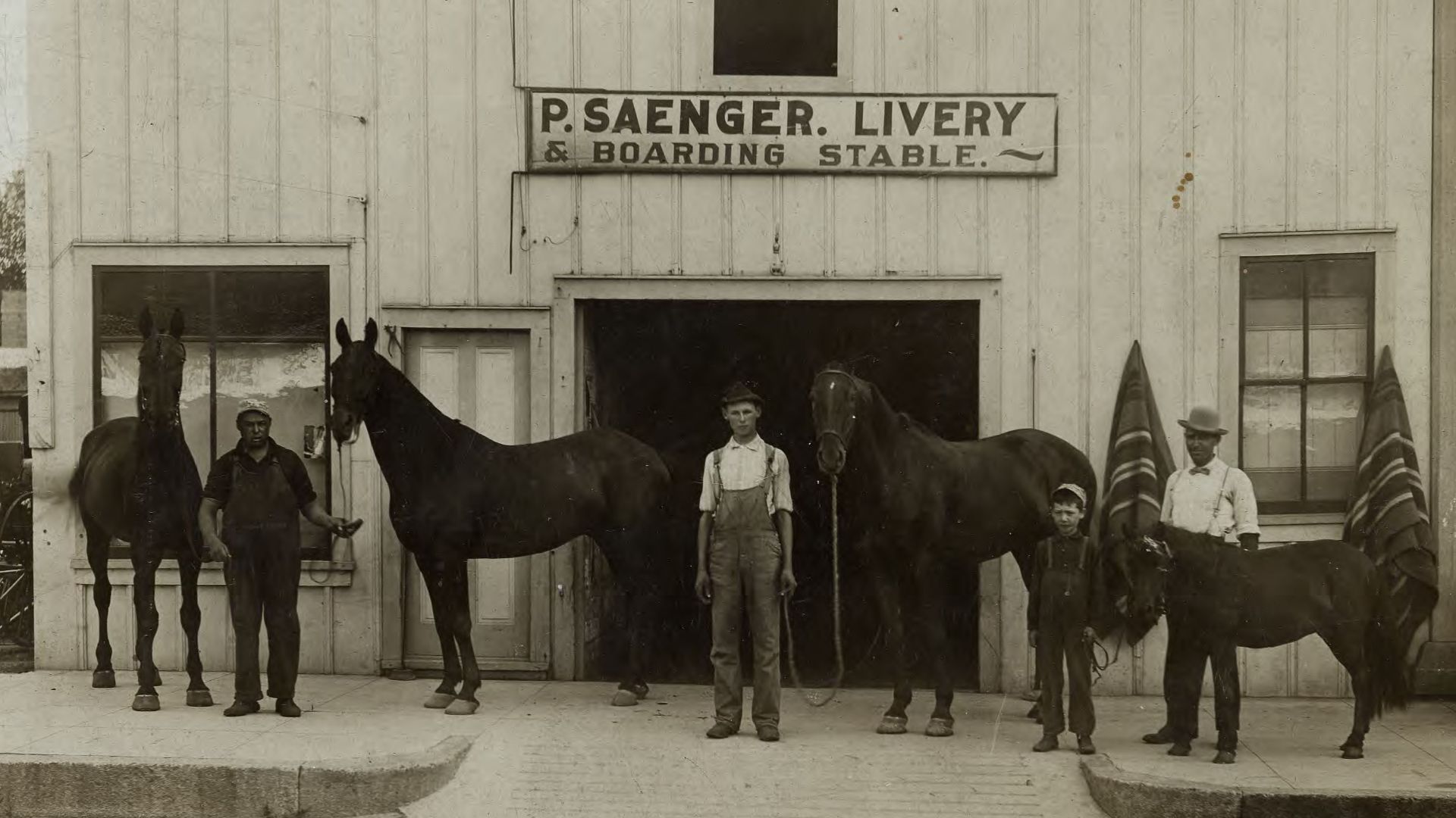 Exterior view of the Saenger Livery Stable with men, a child, three horses and a pony.