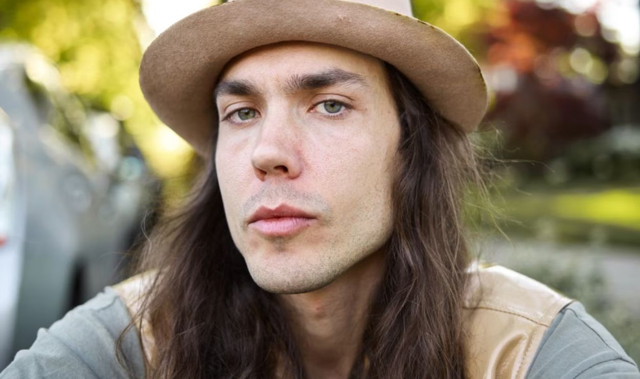 A long-haired man wearing a hat poses confidently outdoors on a sunny day.