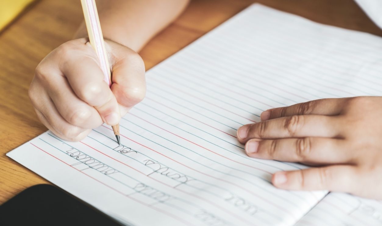 The kid's hand is practicing writing English cursive handwriting sentences in a notebook with a pencil. Cursive handwriting practice. Kindergarten