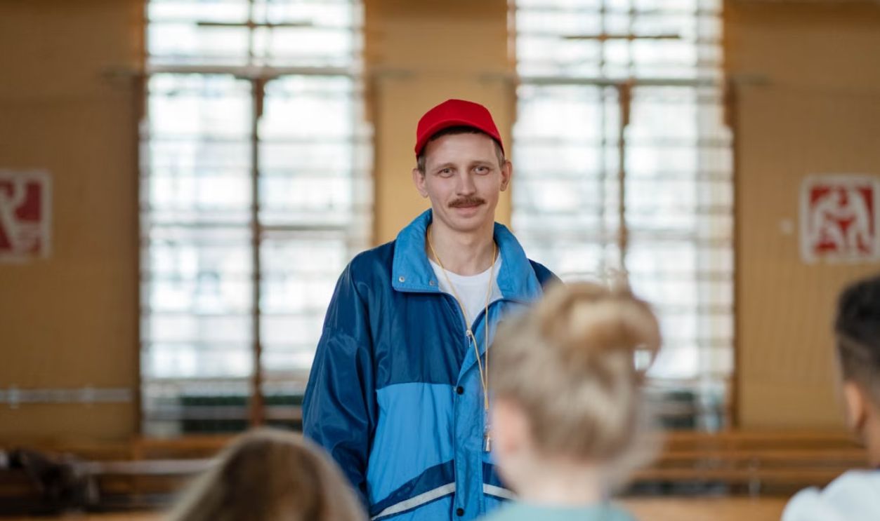 Male coach in a gym engaging with a group of children, wearing casual sportswear.