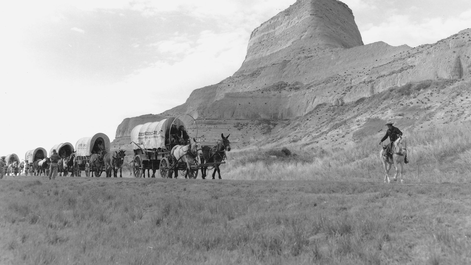 Scope and content:  Mounted man leading wagon train past rock formations.