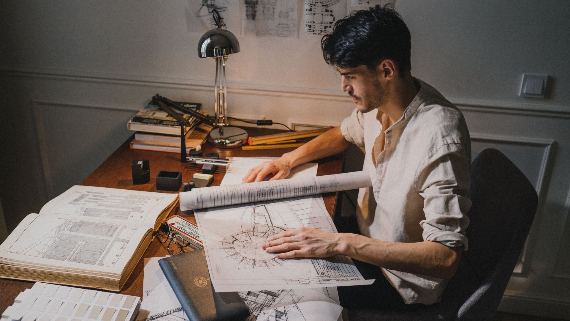 Young architect at a desk working on blueprint designs under lamp light.