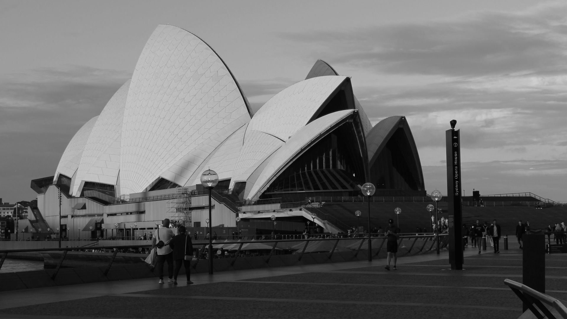 Black and white view of Sydney Opera House at dusk with people walking along the promenade.