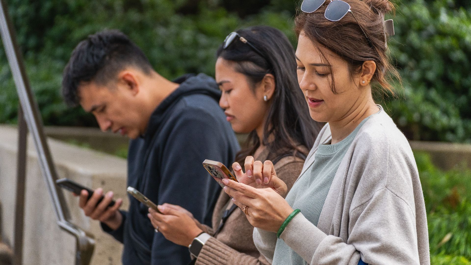 Three young adults focused on their smartphones while sitting outdoors during the day.