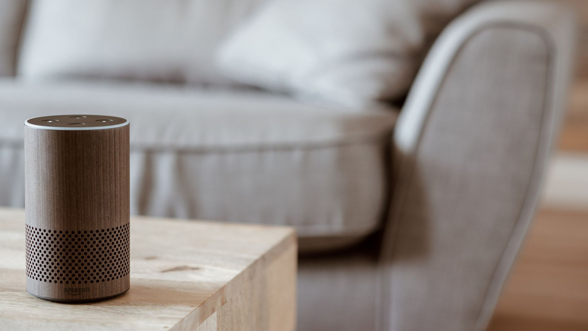 A modern smart speaker sits on a wooden table in a cozy, minimalist living room setting.