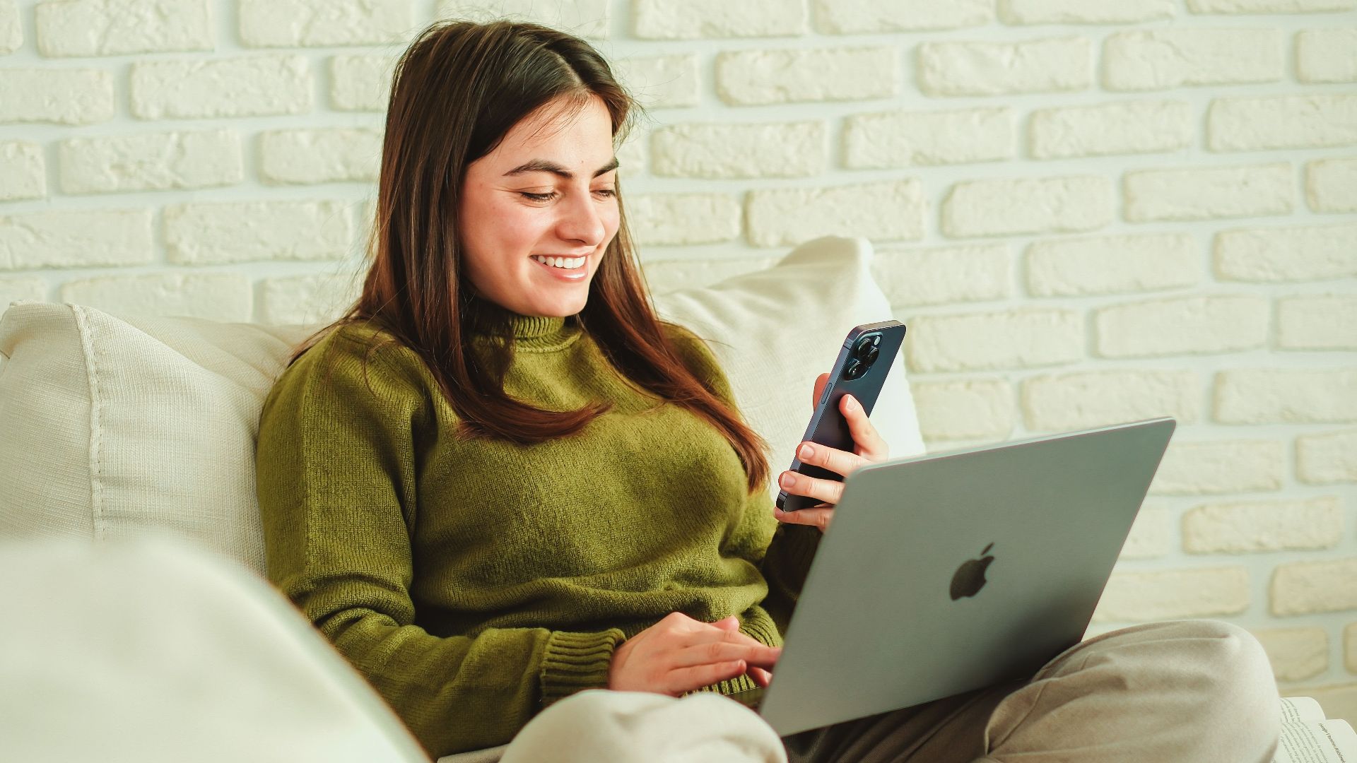Young woman smiling while working on a laptop and phone, seated comfortably indoors.