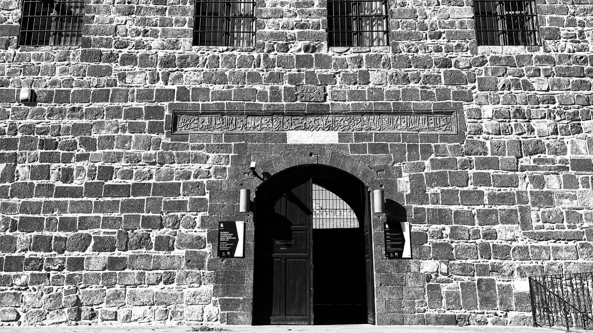 Black and white photo of a historic stone facade with arched doorway and barred windows.