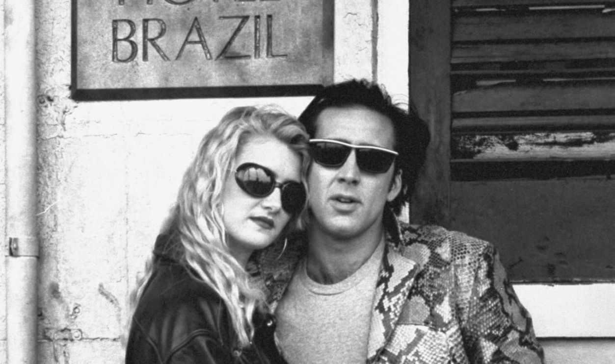 Actor Nicolas Cage wearing a snakeskin sport coat, posing w. actress Laura Dern, both wearing sunglasses, in front of Hotel Brazil on location during filming of movie 'Wild at Heart'.
