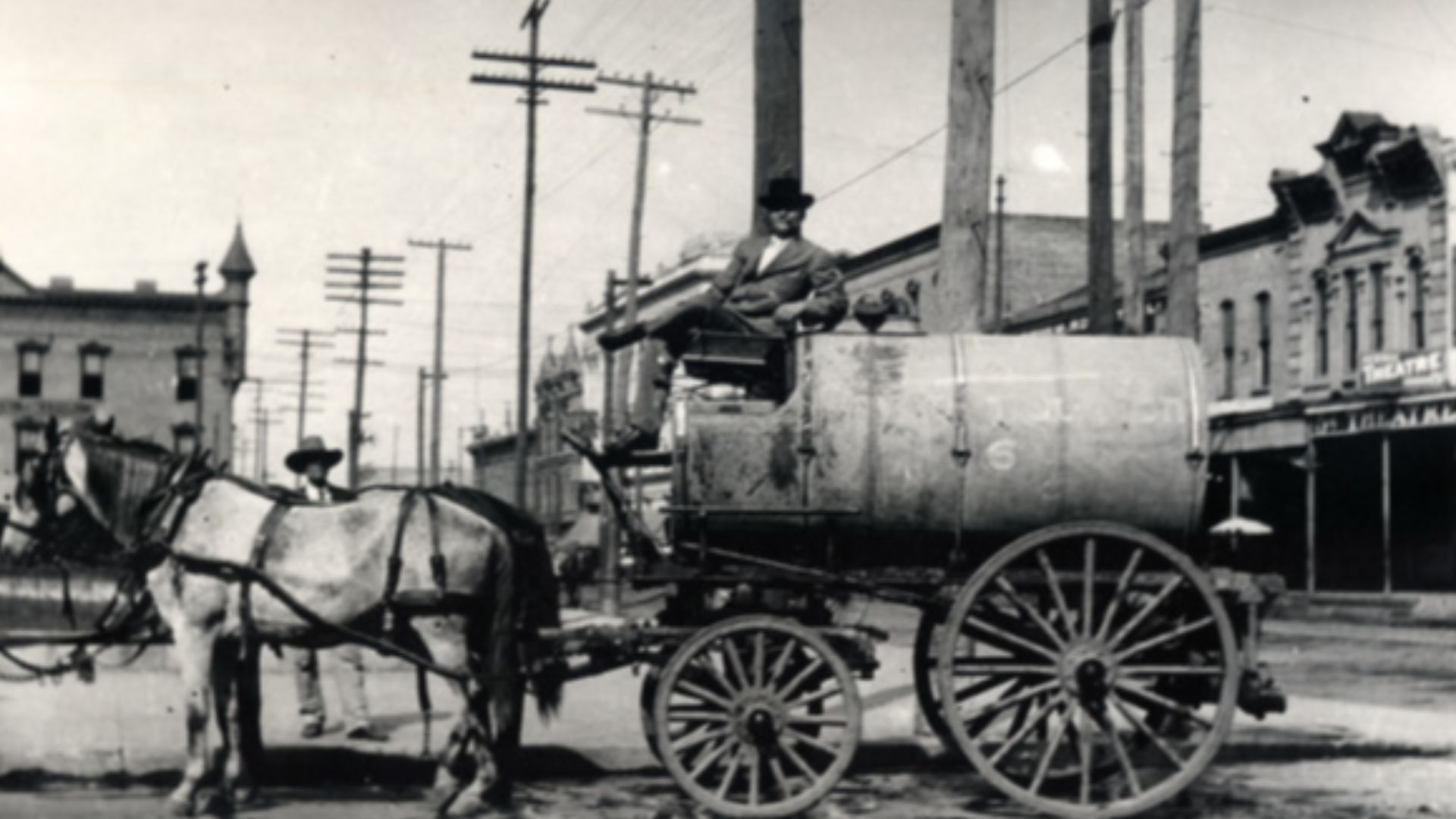 An unidentified man poses in the driver's seat of a horse-drawn water wagon. A second unidentified man stands on the sidewalk. A row of buildings line the street in the background.