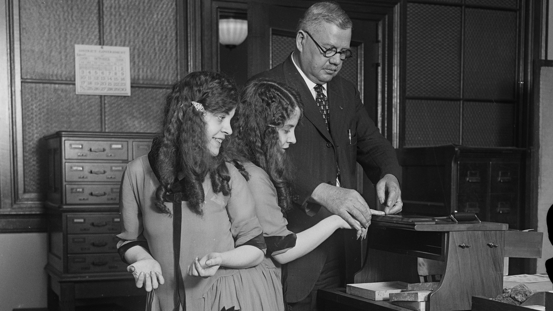 Title: Fingerprinted. Fred Sandberg, District police fingerprint expert, tells Daisy and Violet Hilton,