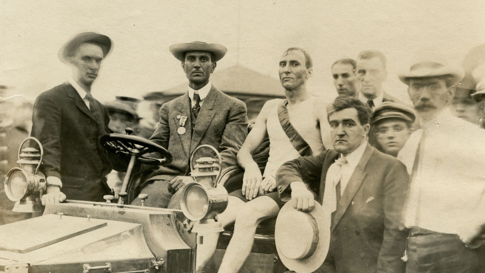Thomas Hicks, winner of Marathon Race, sitting in an open car surrounded by race officials and other men.