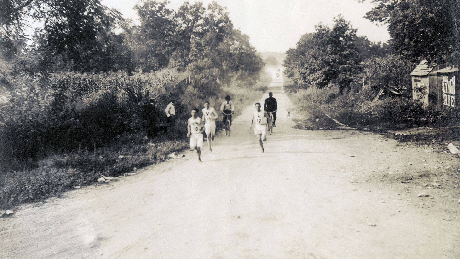 Sam Mellor, Edward Carr, and Michael Spring passing the six mile mark, along a dirt road with judges standing to the side of the road. You can see a corn field and farm out building along the sides of the road.Title: Sam Mellor, Edward Carr, and Michael S