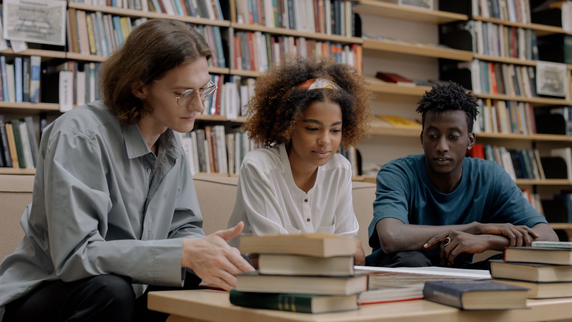 Three students studying together in a library, surrounded by books and learning materials.