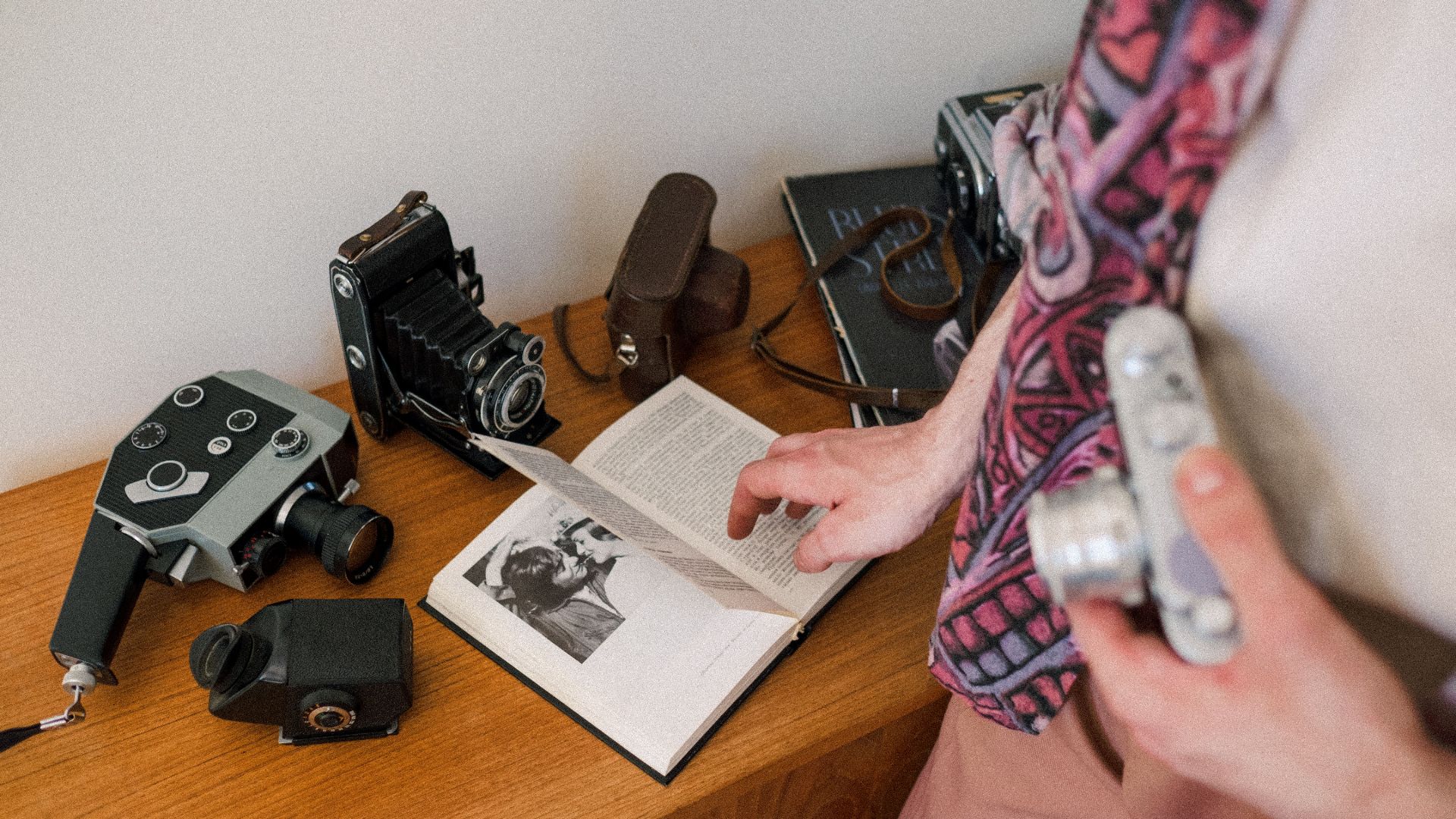 An adult reads a book on a wooden table surrounded by vintage cameras, evoking a passion for classic photography.