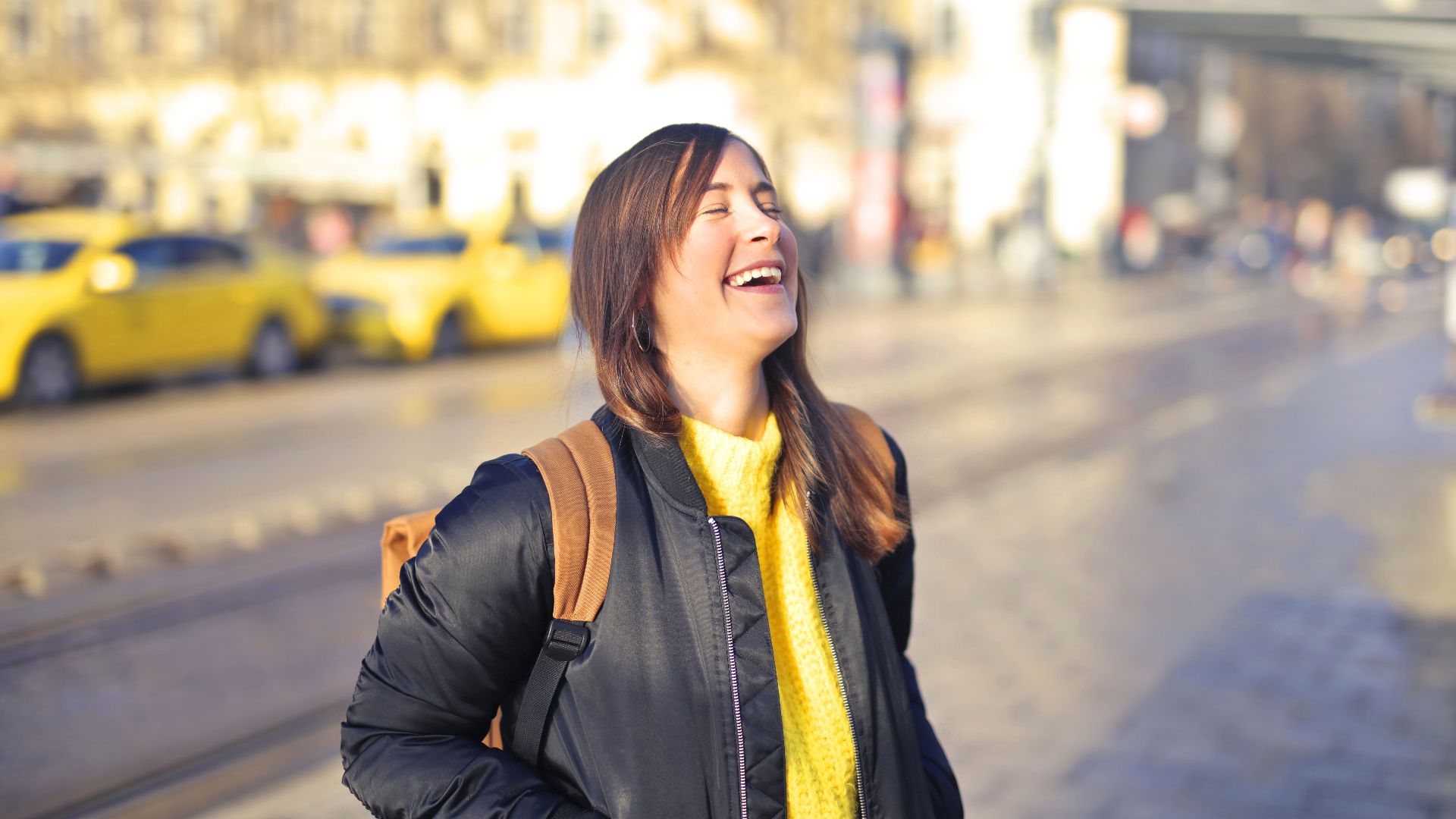 A happy woman in a black jacket laughs on a sunlit city street with yellow taxis.