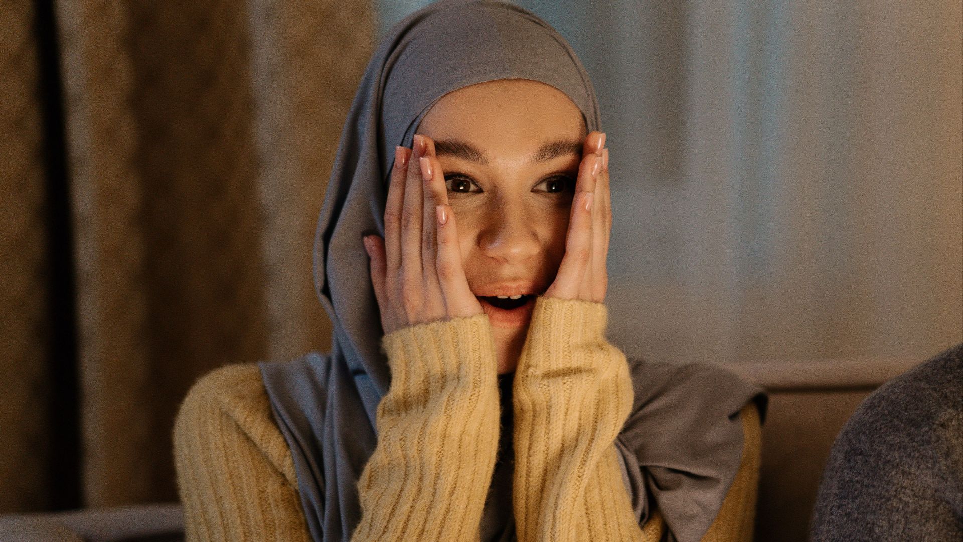 Excited Muslim woman in hijab expressing joy indoors with hands on face.