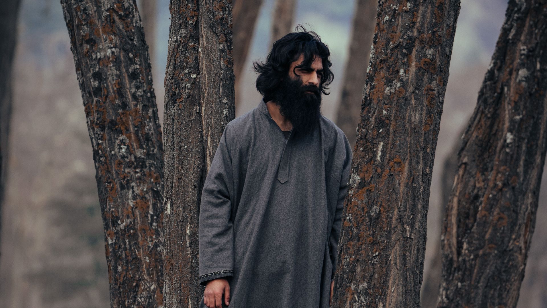 Bearded man in traditional attire standing among tall trees in a forest.
