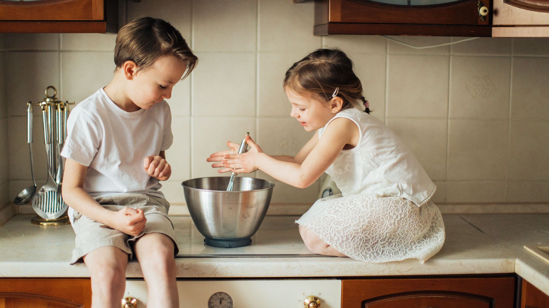 Two kids enjoy baking together on a kitchen counter with a whisk and bowl.
