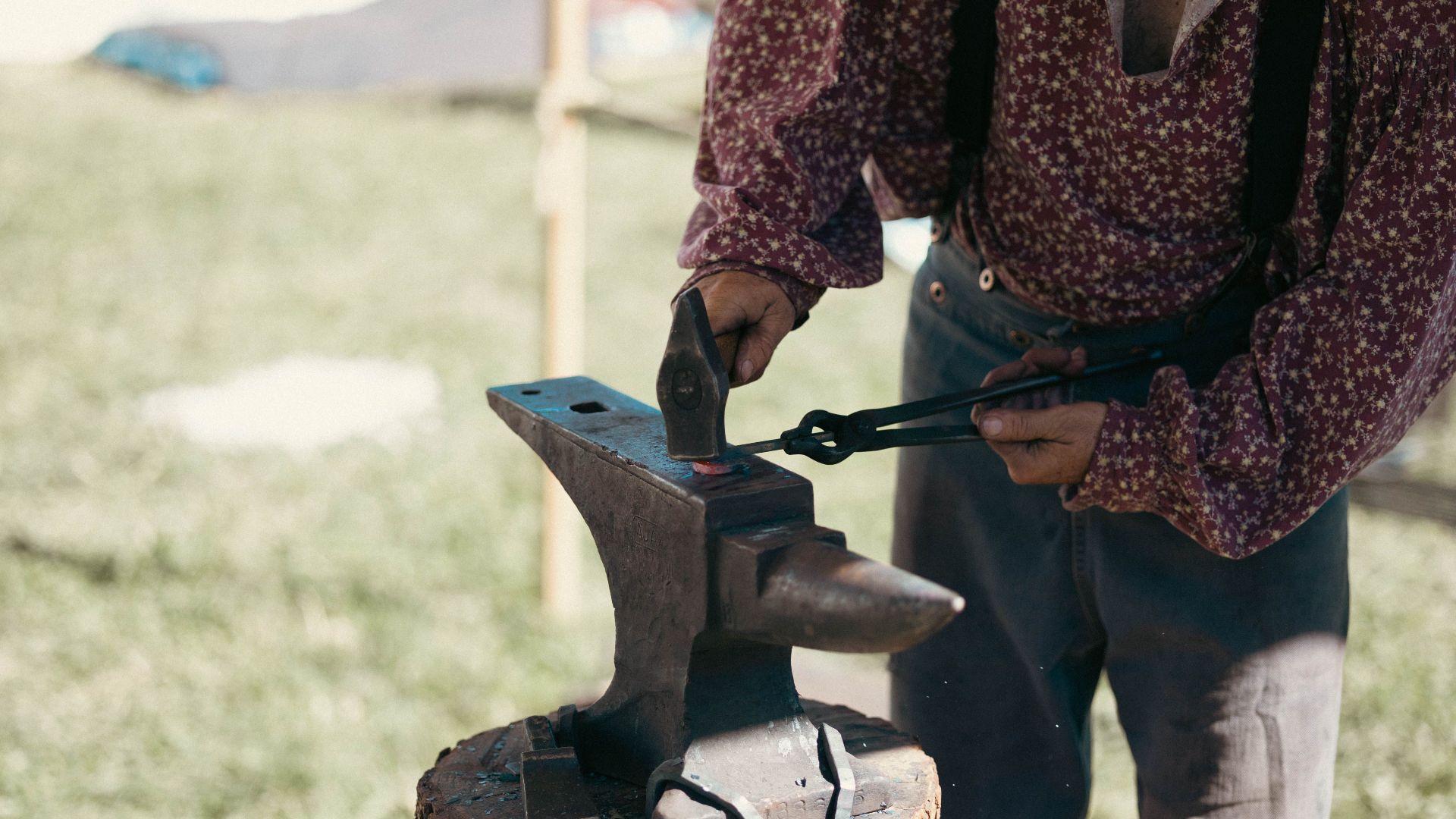 A blacksmith in period clothing is forging metal on an anvil outdoors with tongs and hammer.