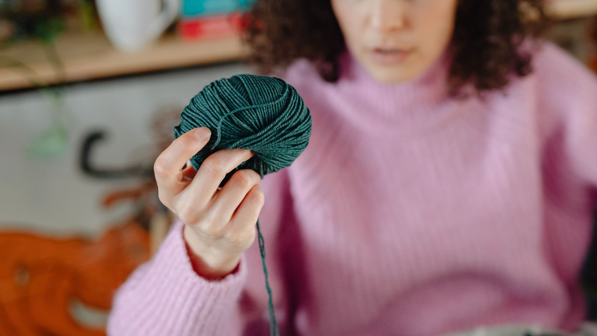 Close-up of a woman holding a green yarn roll, wearing a pink sweater in a cozy indoor setting.