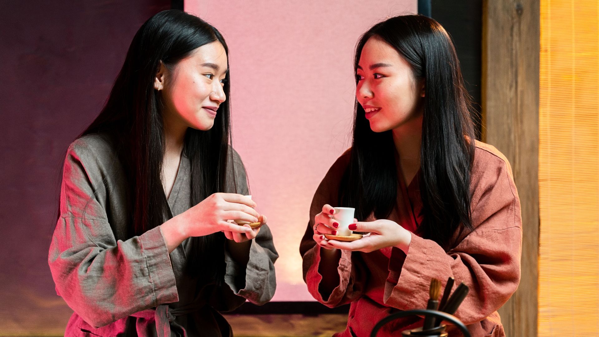 Two young women in warm attire enjoying a cup of tea indoors, sharing smiles.