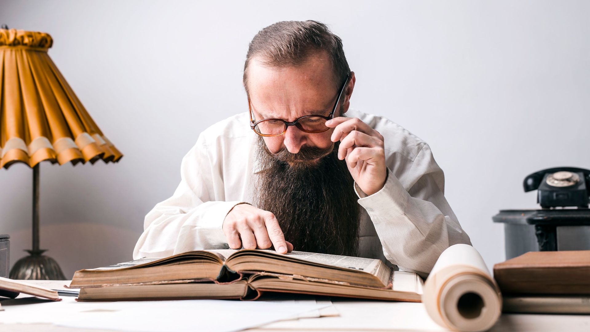 Bearded man with eyeglasses focused on reading a large book at a vintage desk.