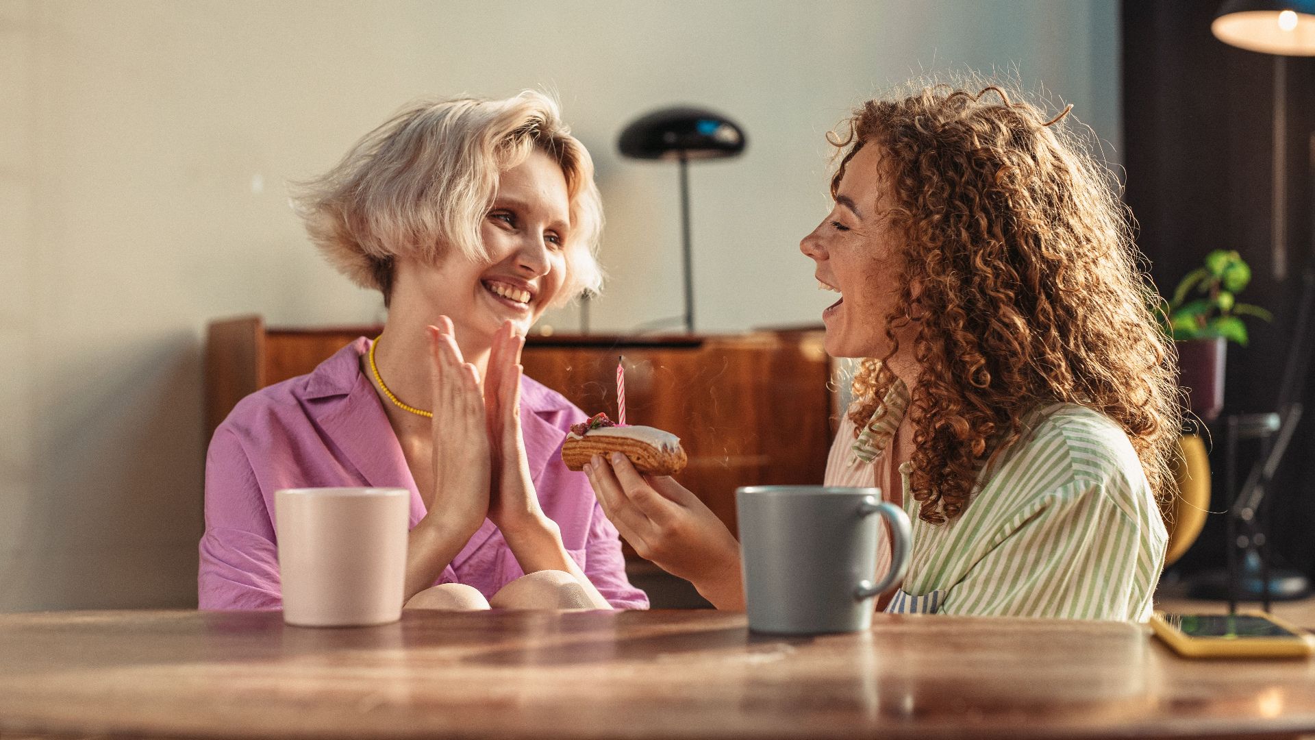 Two friends sharing a joyful moment indoors with coffee and a cake.