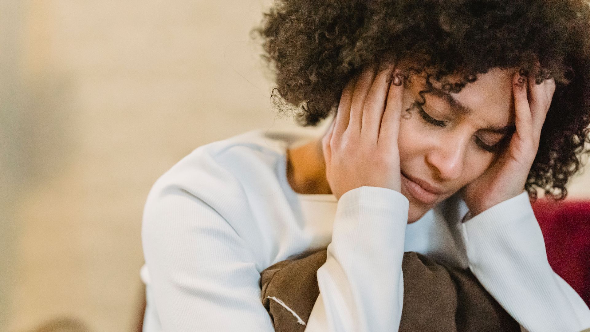 Frustrated African American female with closed eyes touching face while sitting with cushion on couch in room on blurred background