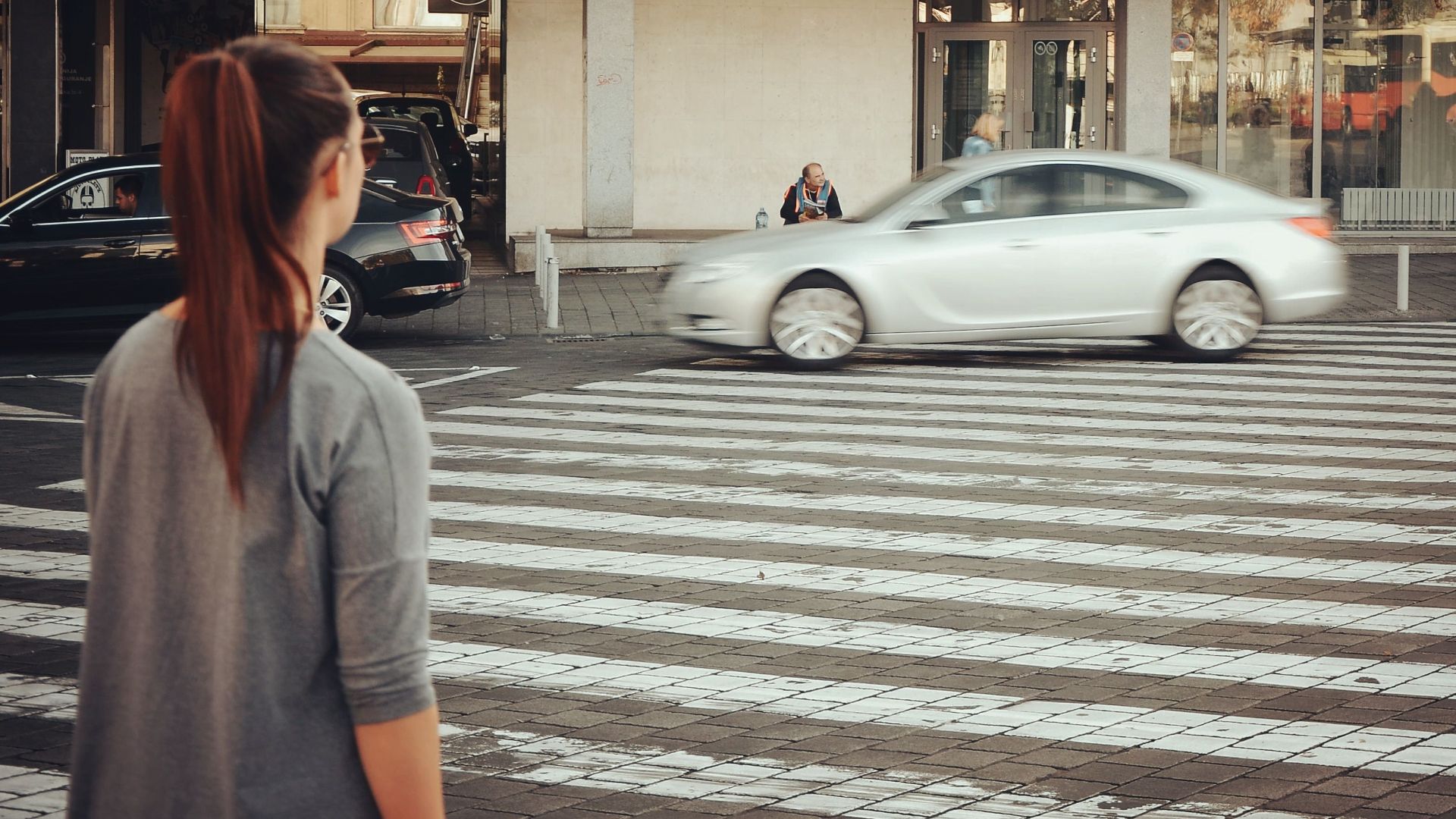 A woman stands at a city pedestrian crossing as cars pass by.