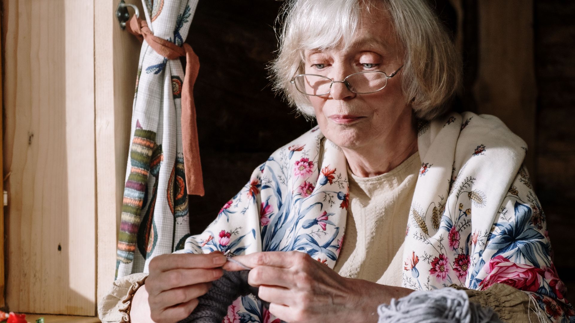 Senior woman knitting with gray yarn near a sunlit window, showcasing peaceful retirement activities.
