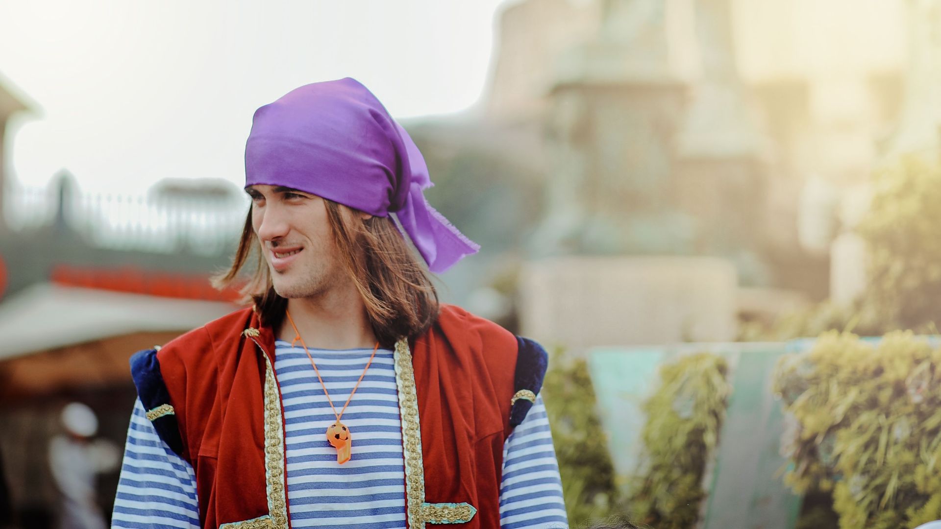 A smiling young man outdoors in a colorful costume and purple bandana.