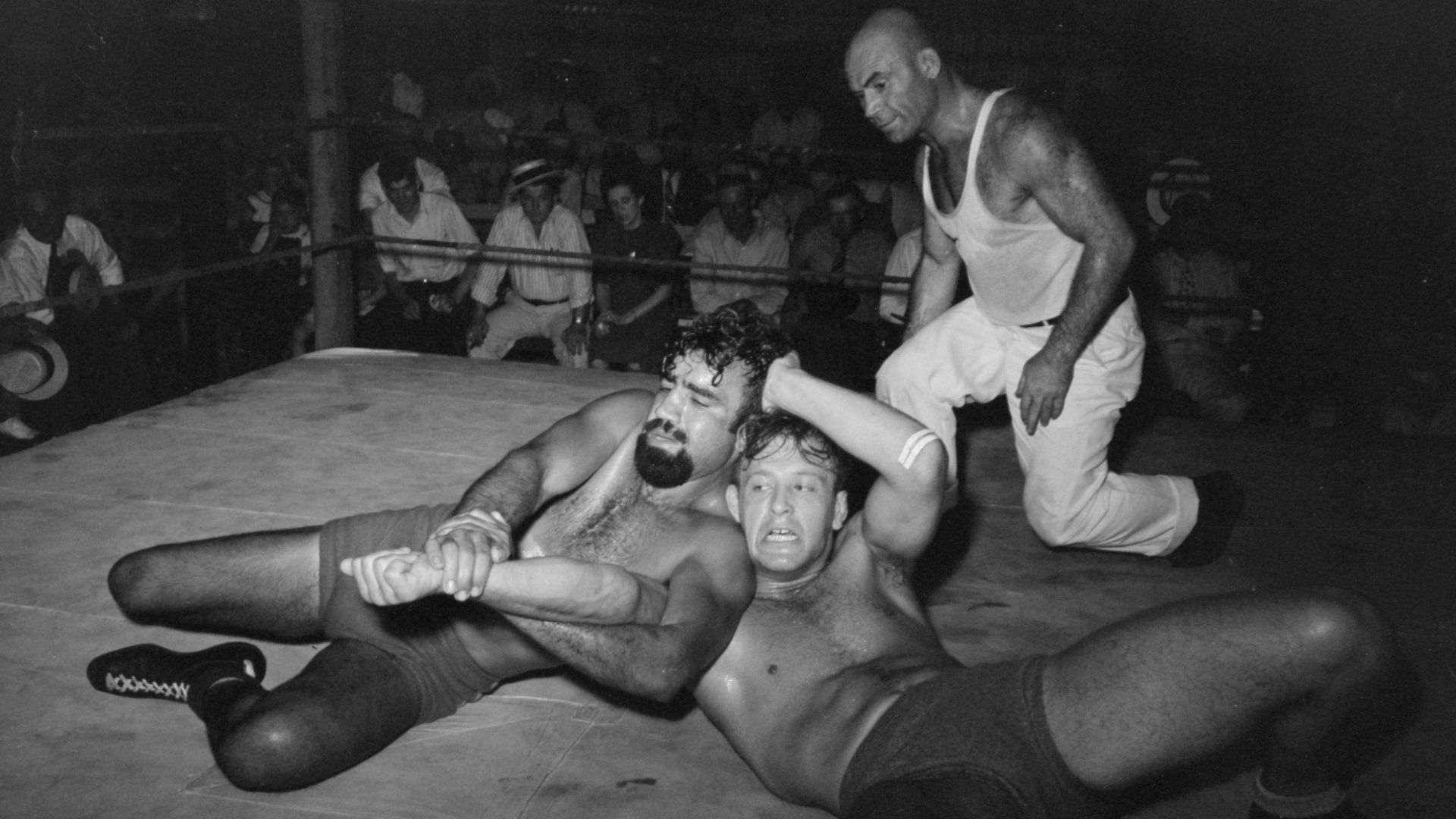 A wrestling match performance in Sikeston, Missouri, May 1938. Two wrestlers grapple while the referee (in white, right) looks on. The bearded wrestler is known as Big Tom Towers. Description on photo: