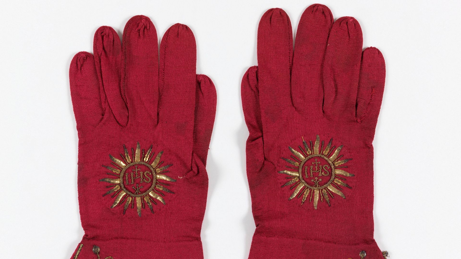 Pair of red Cardinal's gloves with cuffs embroidered in gold metallic yarns and flat gold disks in a vine design with grape clusters. Sacred monogram, IHS, in rayed disk, embroidered in gold on back of each glove. Sewn inside one glove, a handwritten labe