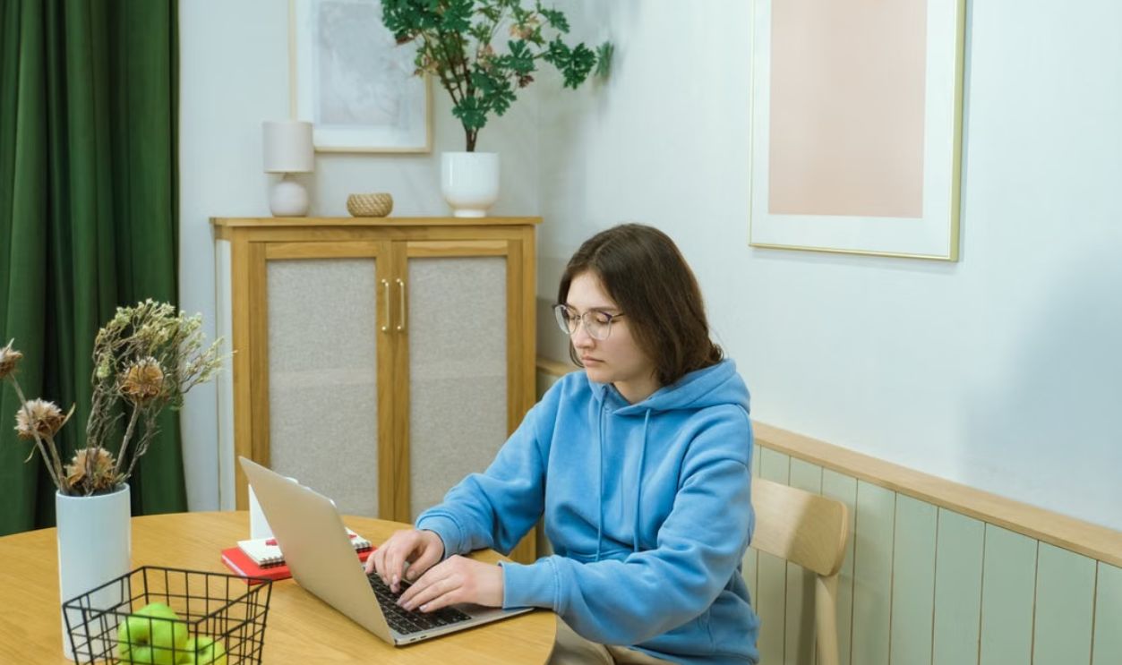 A woman in a blue hoodie works on a laptop at a home office table with green apples and decor