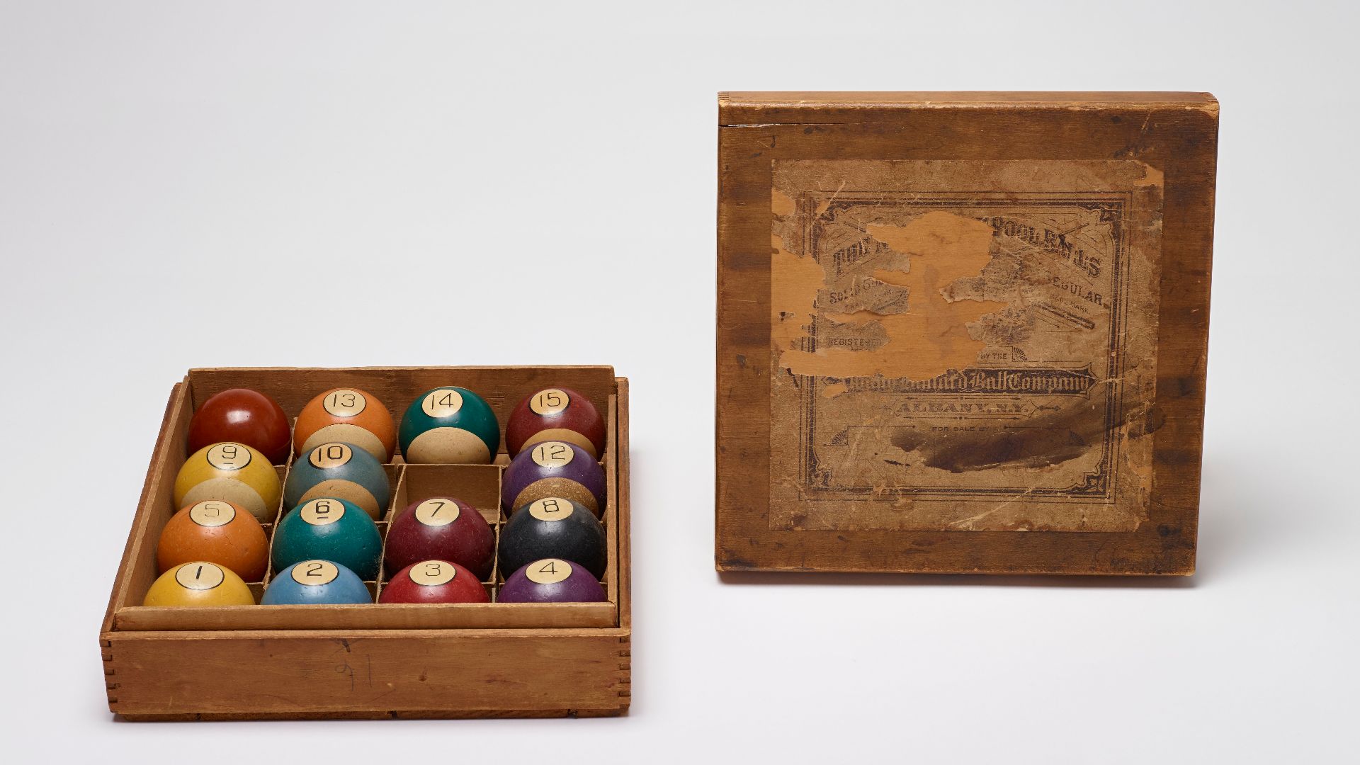 Set of celluloid billiard balls with brown cue ball; balls are divided in bottom section of square wooden box by cardboard dividers.