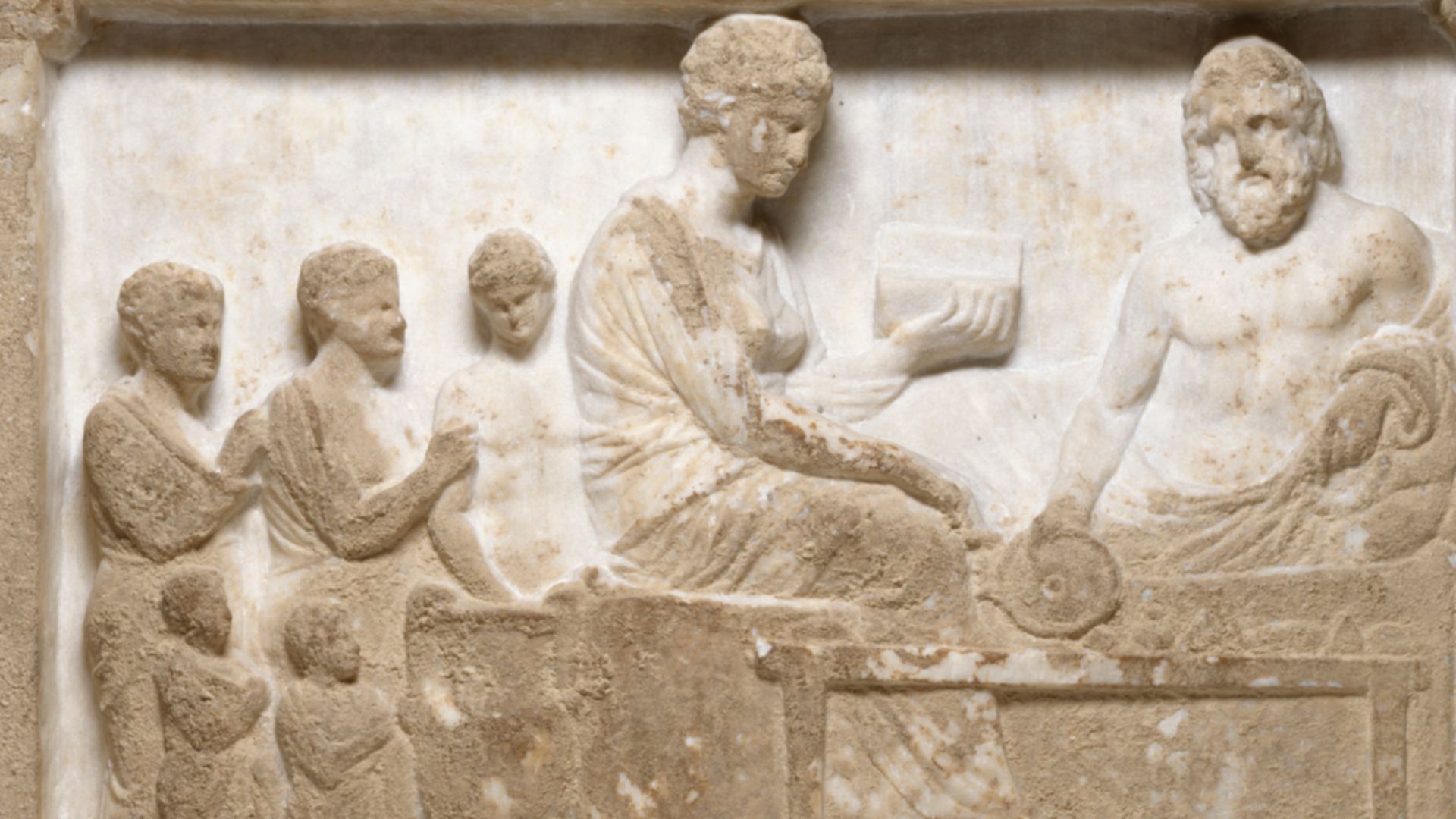 The banquet scene, of Near Eastern origin, was used for Classical funerary reliefs and also as votive offerings to gods. Here, the deceased male reclines at his funerary banquet accompanied by his wife and family. The large vessel holds wine; on the table