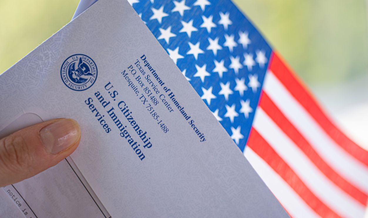 Washington, DC, USA - September, 16, 2019: Letter (Envelope) from USCIS ans flag of USA in hand on green blurred background. Diagonal Close up view.