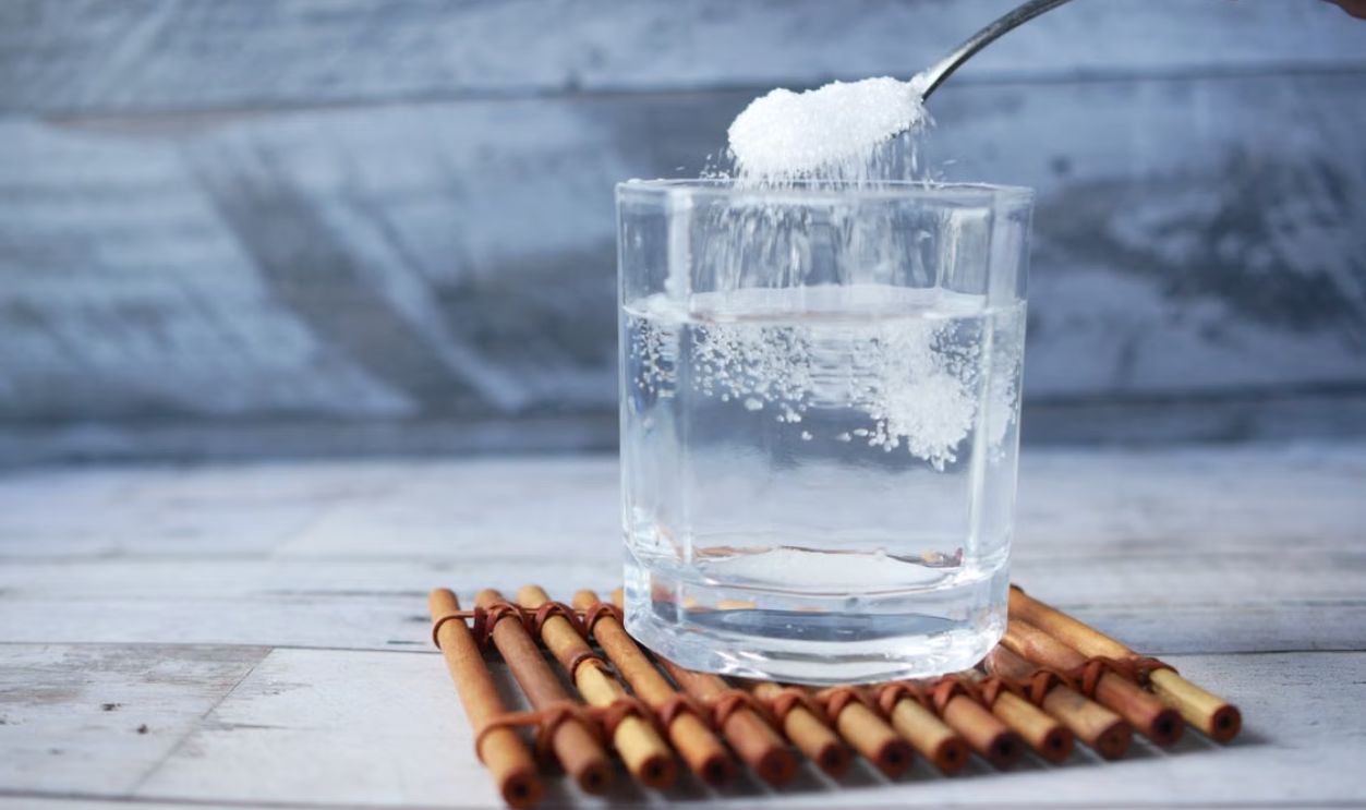 A spoonful of sugar being added to a glass of water on a wooden mat.