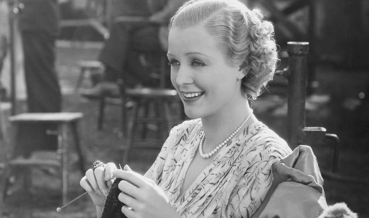 (Original Caption) In a sunlit spot during an outdoor shooting operation of Professional Soldier, Gloria Stuart pauses in her knitting to smile at something occurring before the cameras.