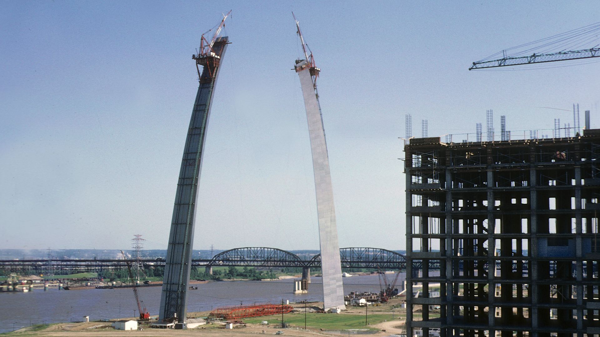 The Gateway Arch in St. Louis, MO, under construction in June 1965, before the truss used to support the legs of the arch is lifted into place. The truss can be seen on the ground underneath the arch. MacArthur Bridge is visible in the background.
