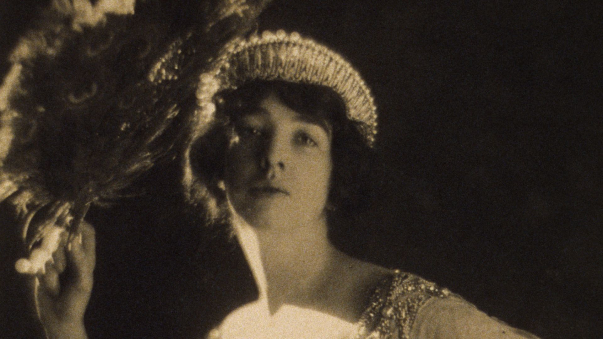 Gertrude Vanderbilt Whitney wearing a jeweled gown and tiara and holding a peacock feather fan.