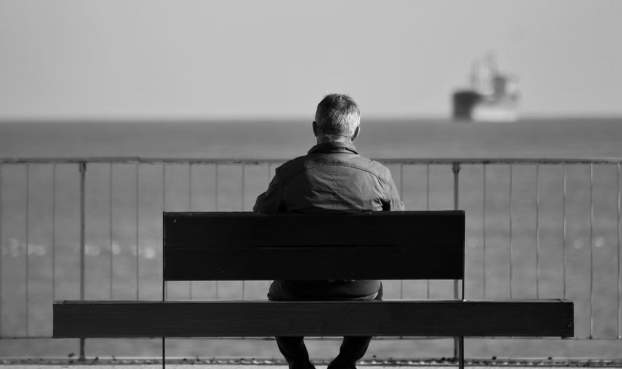 Man sits on bench by the sea, gazing at distant ship, evoking solitude